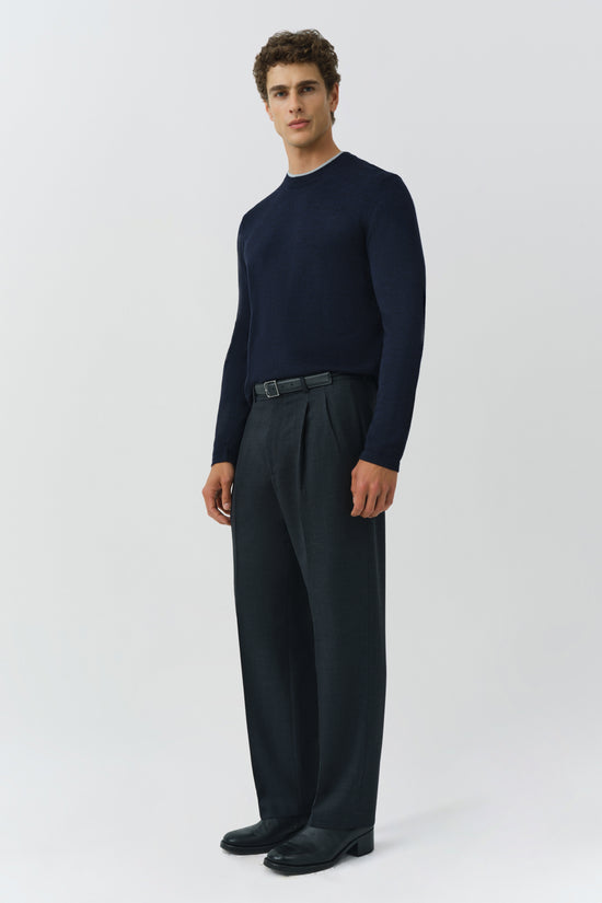 Man wearing dark pleated trousers with a belt and navy crewneck sweater.