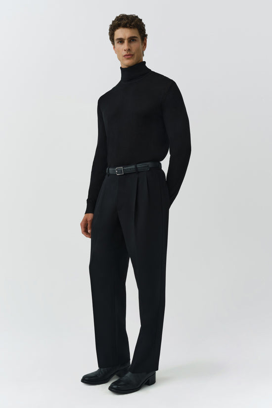 Man wearing black turtleneck with black trousers and leather belt against neutral background.