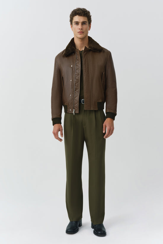 Brown leather jacket with dark fur collar, snap button closure, and quilted lining worn with olive pants.