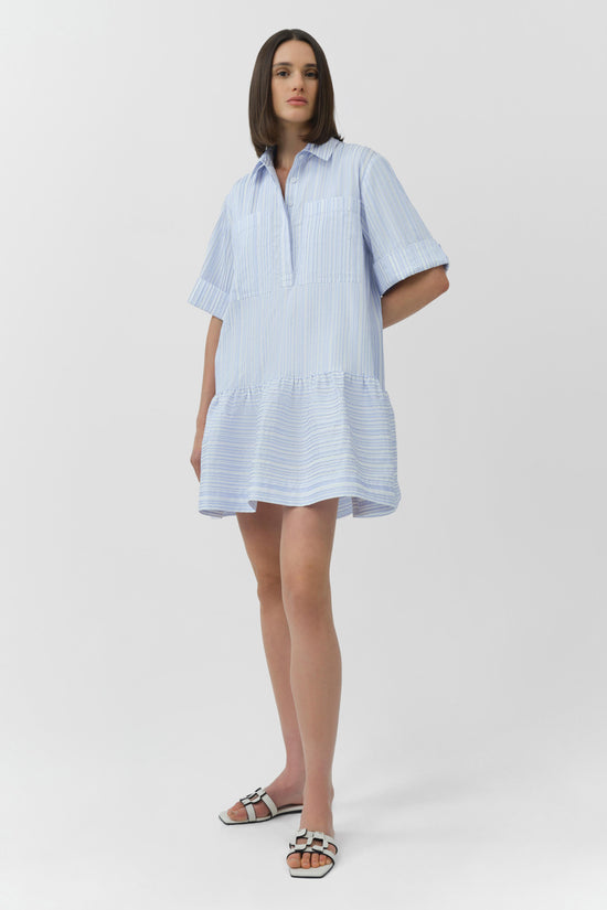 Light blue striped short sleeve shirt dress with button front and gathered hem, worn with white slide sandals.