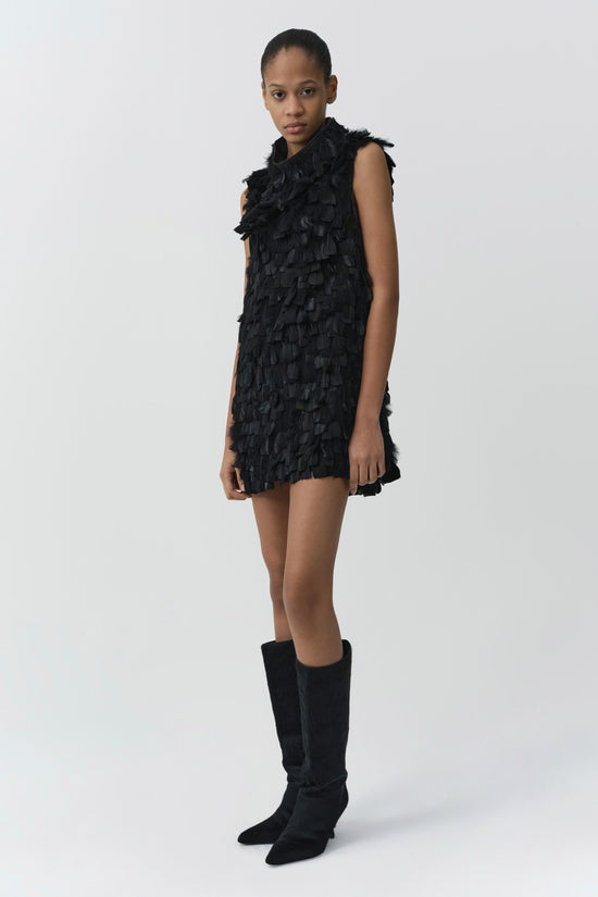 Black sleeveless mini dress with layered feather detailing throughout, styled with black knee-high boots.