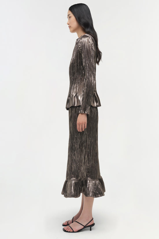 Woman wearing a silver metallic pleated long-sleeve top with ruffled cuffs and hem, paired with black strappy heels.