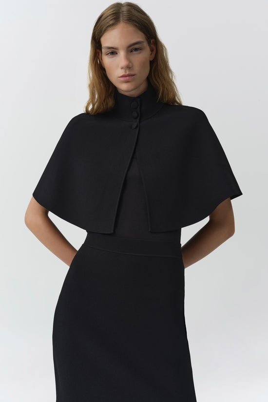 Black knit cape with high neckline and button closure worn over a matching black dress.