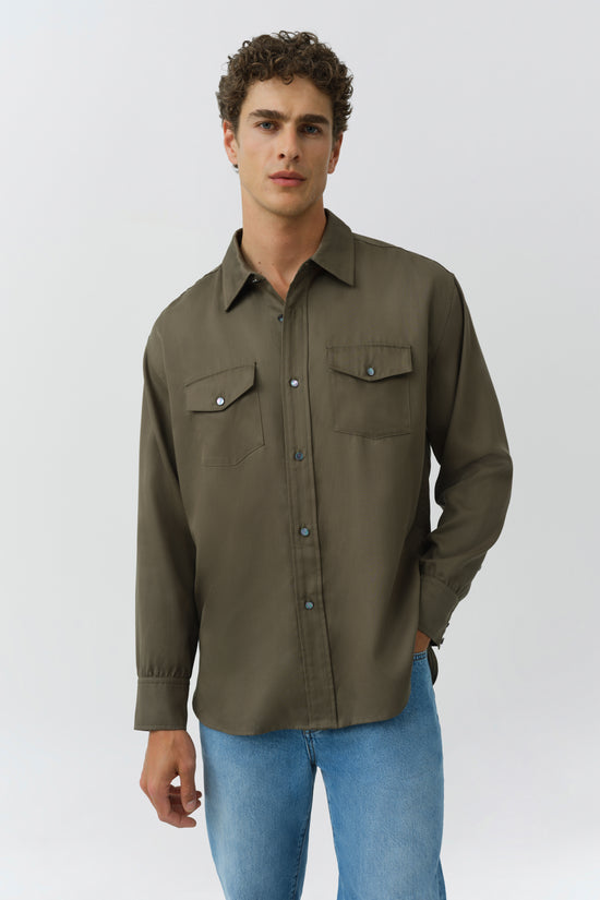 Olive green button-up workwear shirt with chest pockets and long sleeves on model wearing blue jeans.