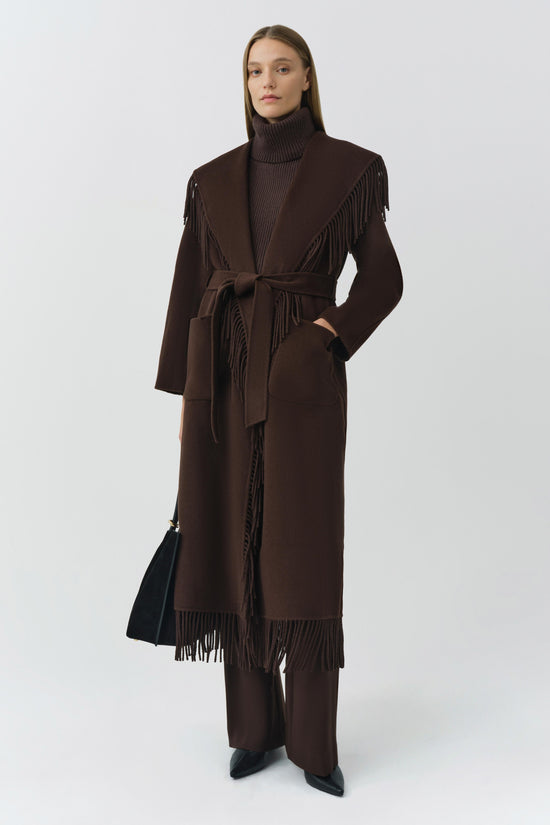 Brown wool coat with fringe trim on shoulders and hem, tie waist belt, and notch lapels worn over turtleneck.