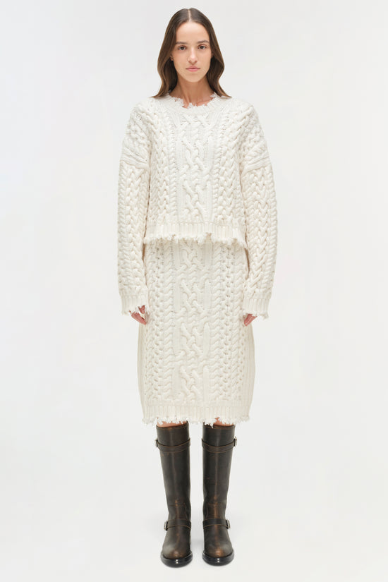 Cream cable knit cropped sweater with distressed edges worn with a matching midi skirt and black boots.