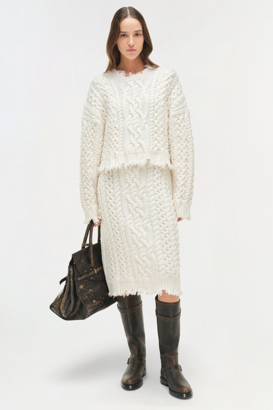 Cream cable-knit cropped sweater with distressed edges and textured pattern worn with black boots.
