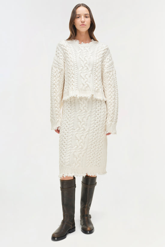 Cream cable knit cropped sweater with distressed edges worn with matching skirt and black boots.