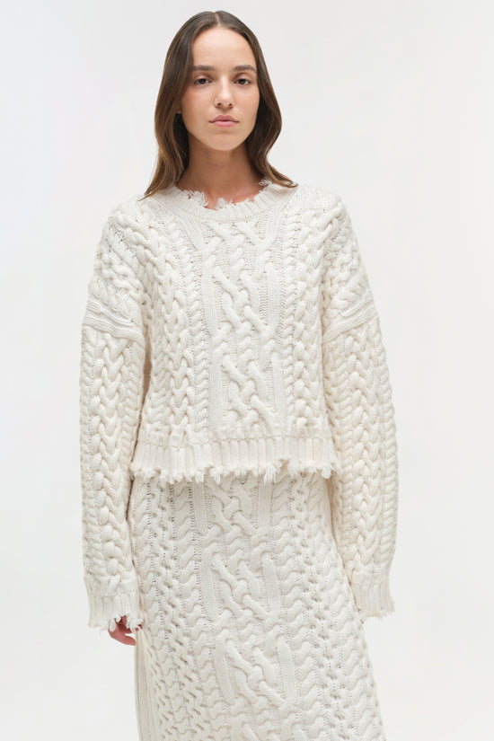 Cream cable knit cropped sweater with distressed edges and frayed hem worn with matching skirt.