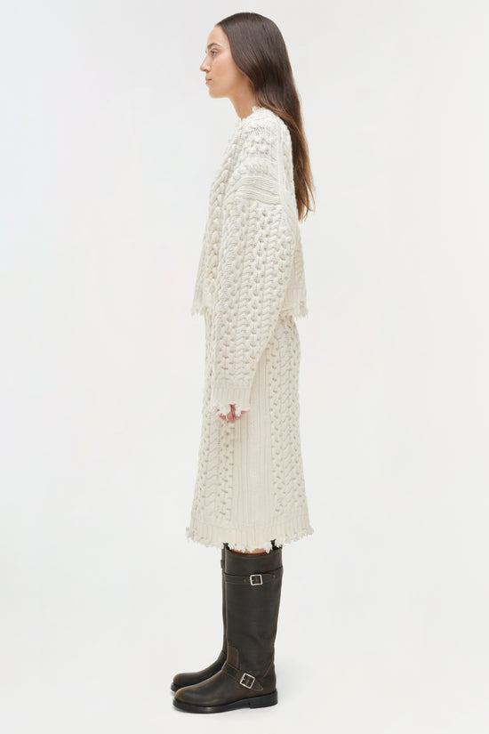 Woman wearing a cream cable-knit cropped sweater with distressed details, styled with black knee-high boots.