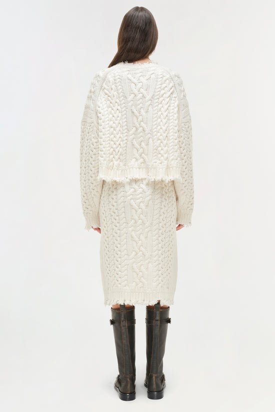 Cream cable knit cropped sweater with raw edges worn over a matching midi skirt, back view.