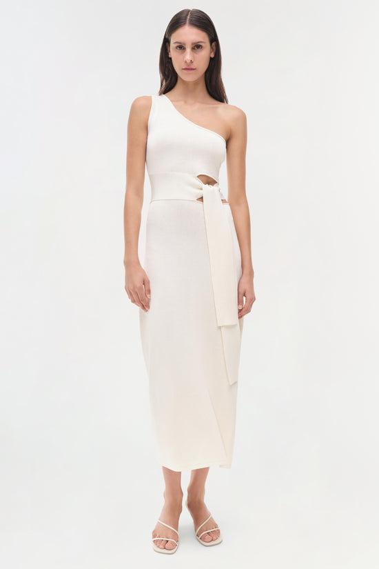 Woman wearing cream one-shoulder dress with cutout waist and tie detail, paired with white strappy sandals.