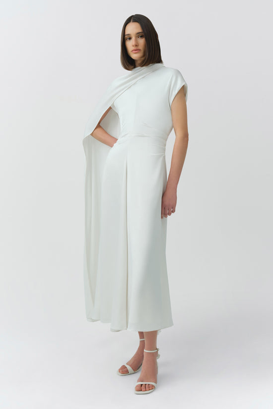 Cream satin midi dress with asymmetrical cape drape and cutout at waist, shown on model with white sandals.