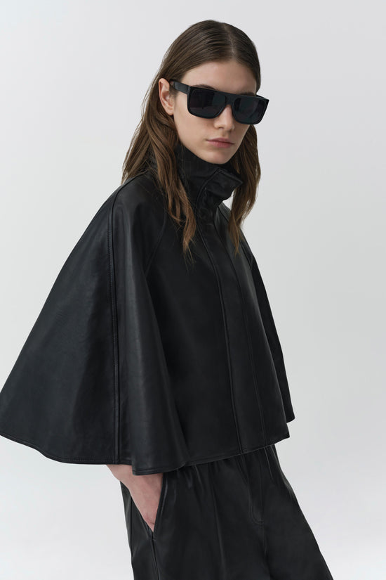 Black leather cape jacket with front zipper and collar, worn over matching pants.
