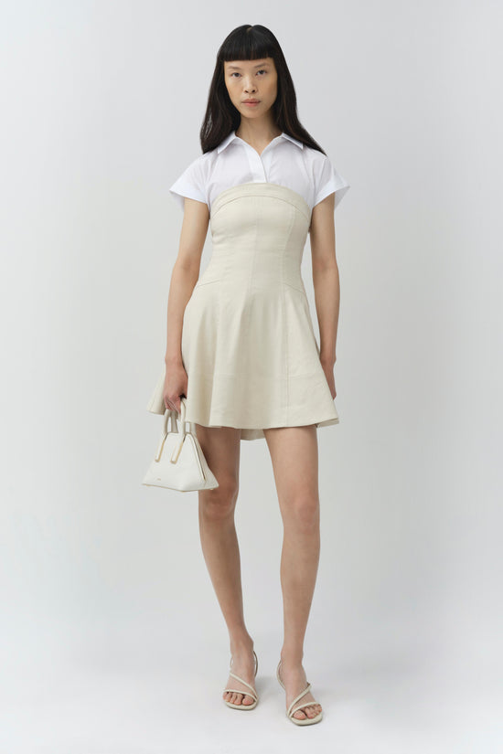 Woman wearing white short-sleeved collared shirt over cream-colored sleeveless A-line mini dress, holding matching handbag.