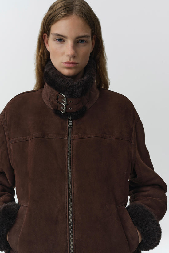 Brown suede jacket with black shearling collar, cuffs, and pocket trim, featuring a front zip closure and buckle neck detail.