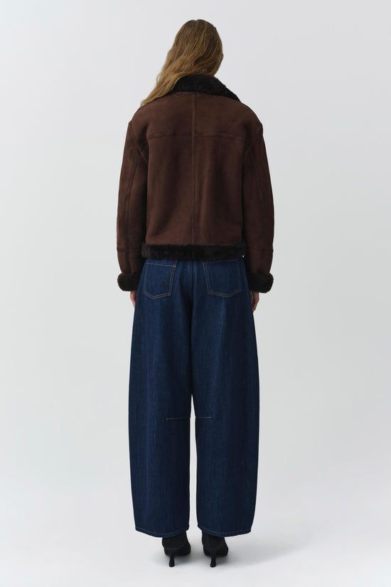 Brown suede jacket with black shearling collar and cuffs, worn with dark blue jeans.