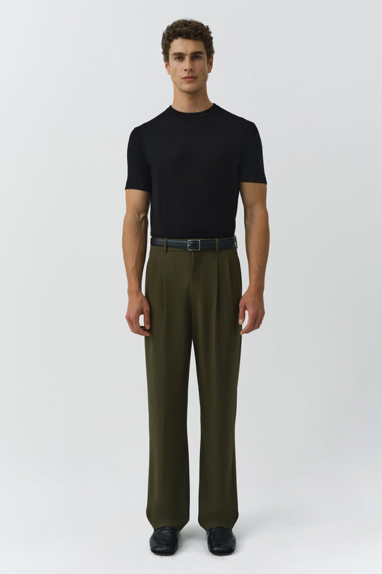 Man wearing black t-shirt and olive green pleated trousers with belt and black shoes.