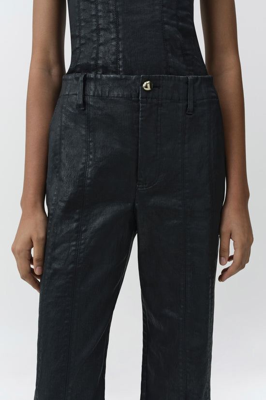 Dark coated denim trousers with high waist, straight leg, and gold-tone button closure on model.