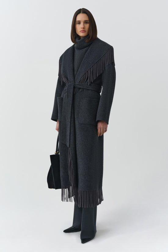 Charcoal wool coat with fringe trim on shoulders and hem, belted waist, worn with turtleneck and black boots.