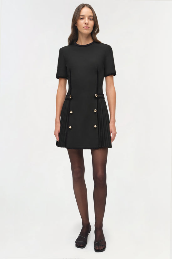 Black mini dress with short sleeves, gold button accents at waist, and pleated skirt detail.