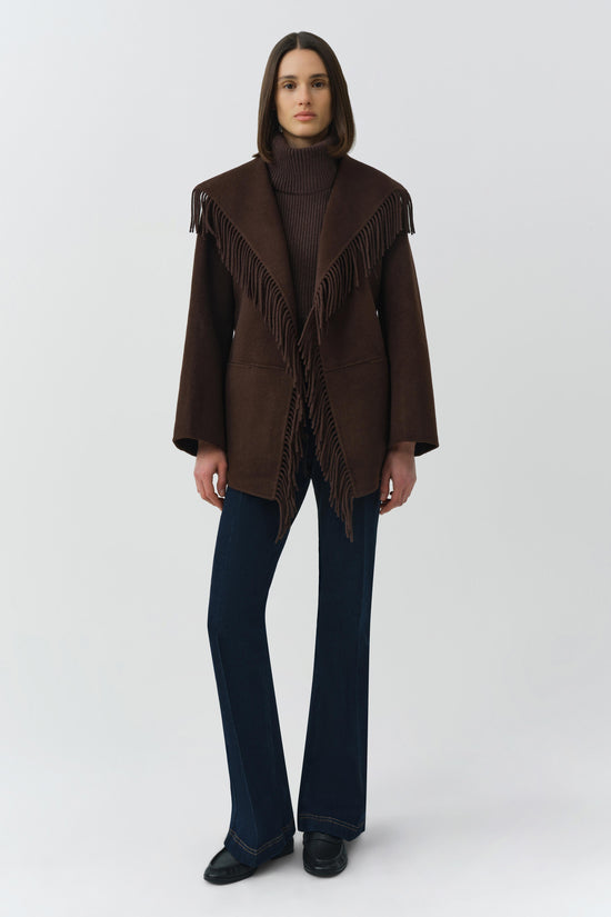 Brown wool jacket with fringe trim on collar and shoulders, worn with dark turtleneck and navy flare jeans.