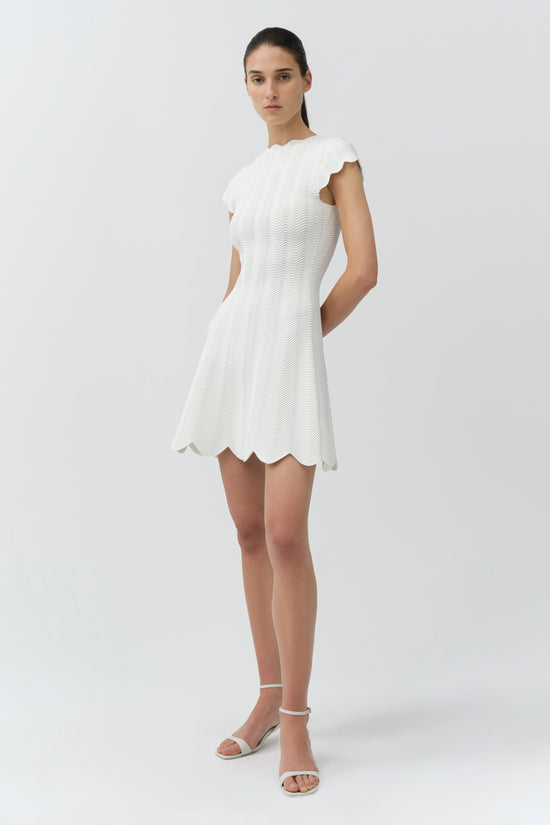 White wavy knit mini dress with short ruffled sleeves and scalloped hem on model with white sandals.