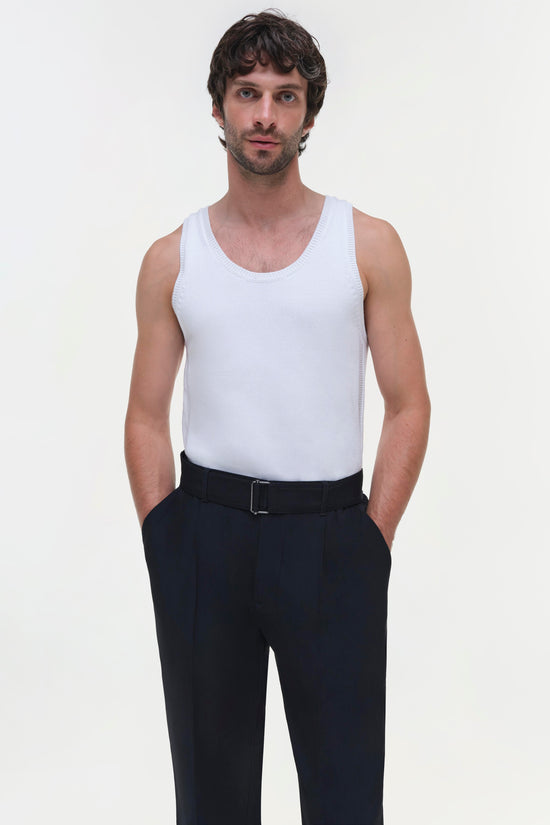 Man wearing black trousers with belt and white sleeveless tank top against neutral background.