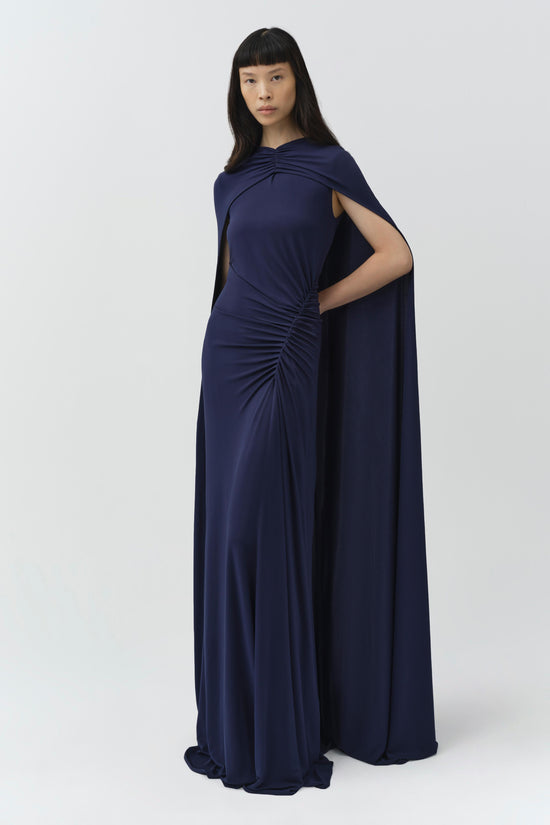 Woman wearing navy draped jersey cape gown with ruched waist detail and floor-length skirt.