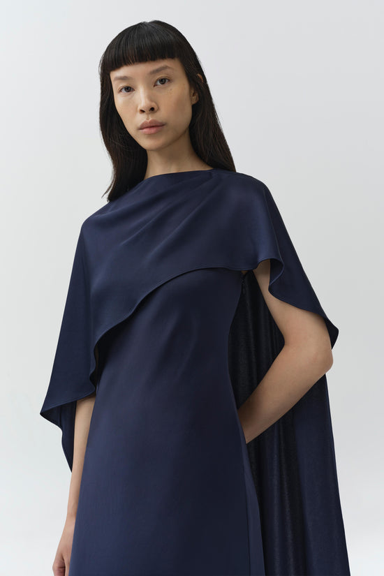 Navy satin cape gown with draped overlay and sleek silhouette on model against neutral background.