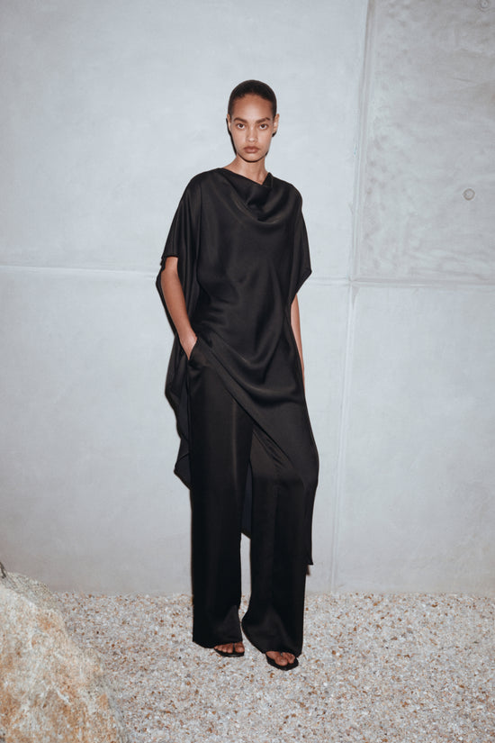 Black satin mini cape dress with draped fabric, cowl neckline, and side pockets on model.