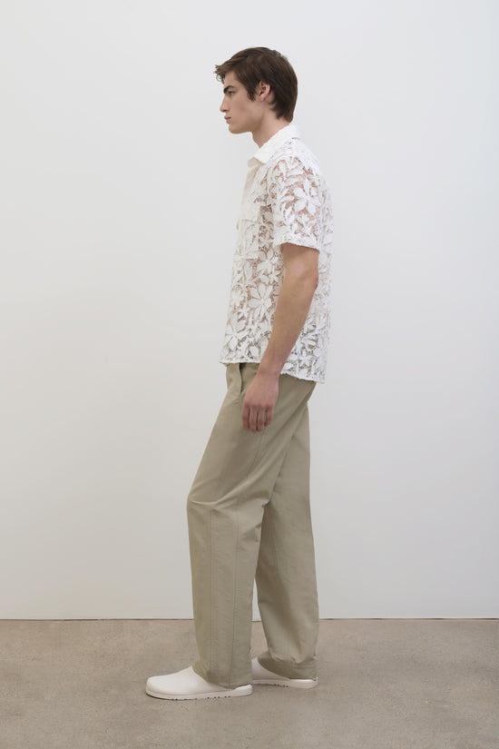 White cotton lace short-sleeve shirt with collar, shown on model wearing beige trousers and white shoes.