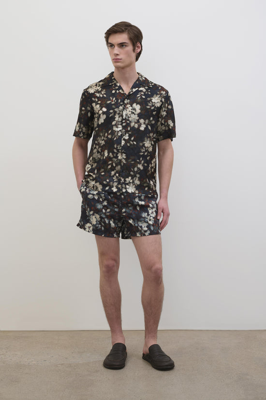 Man wearing black silk shirt with cream floral print and matching shorts, styled with black slip-on shoes.