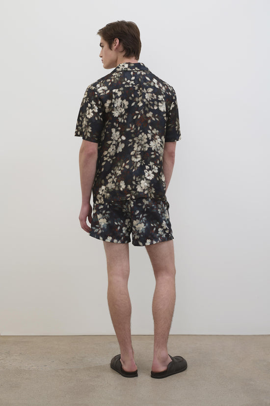 Man wearing black swim shorts with cream and tan floral print, shown from behind with matching floral shirt.