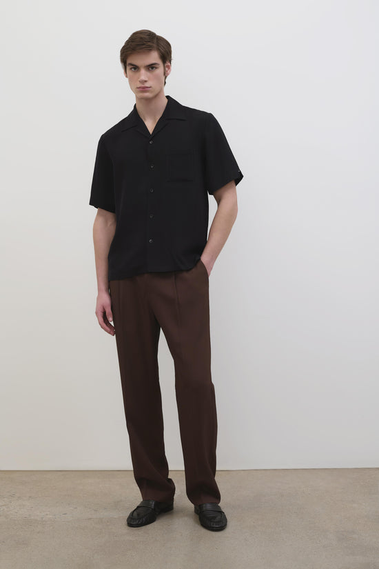 Man wearing black short-sleeve camp shirt with collar and button front, paired with brown trousers and black slippers.
