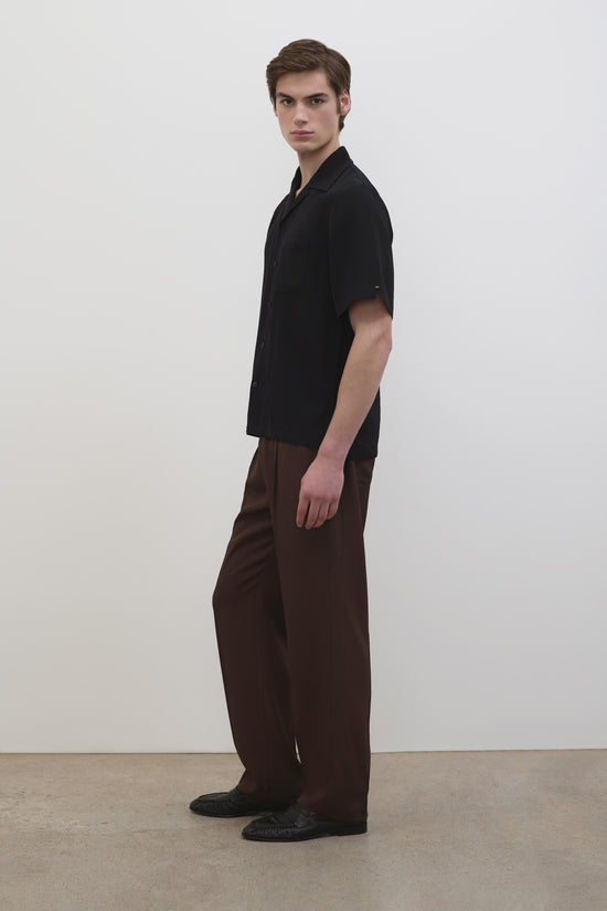 Man wearing black short-sleeve camp shirt with brown trousers and black shoes.