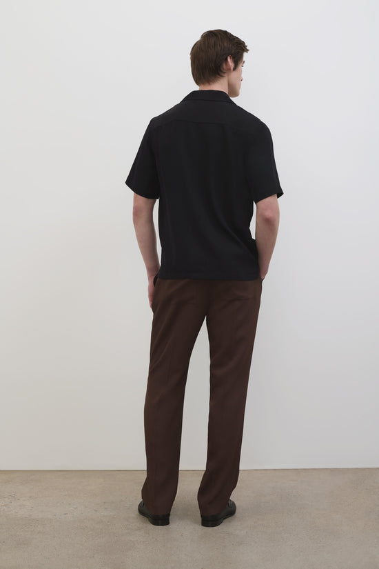 Man wearing black short-sleeve camp shirt with brown pants, shown from behind.