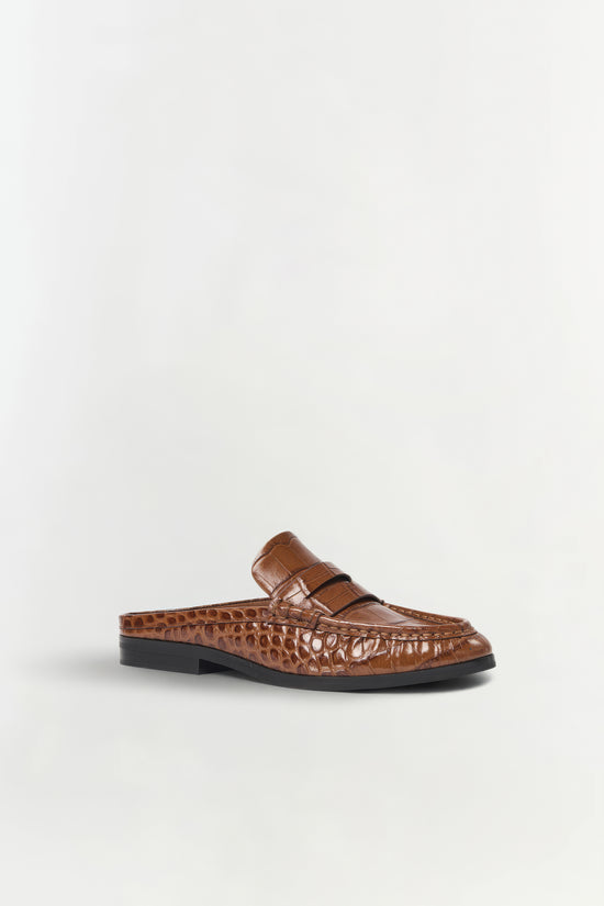 Brown crocodile-textured leather loafer mule with black sole, shown from the side view.
